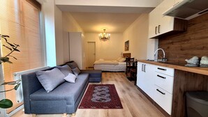 A compact living space with a grey sofa, a small dining table, a kitchen area with wooden cabinets, and a red patterned rug. - KindStay Suites Prague Airport (Středočeský kraj)
