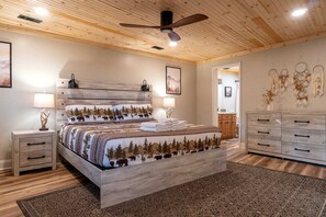 Quarto com teto de madeira, cama com colcha temática de floresta, luminárias de cabeceira, mesa de cabeceira, cômoda e ventilador de teto.