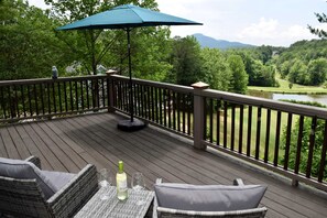 Eine Holzterrasse mit Gartenmöbeln, einem Tisch mit Gläsern und einer Flasche sowie Blick auf einen See und Wald.