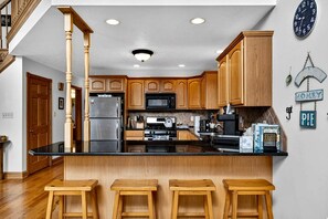 Una cocina con armarios de madera, un refrigerador de acero inoxidable, un microondas y una cafetera. Hay tres taburetes de madera en la encimera.
