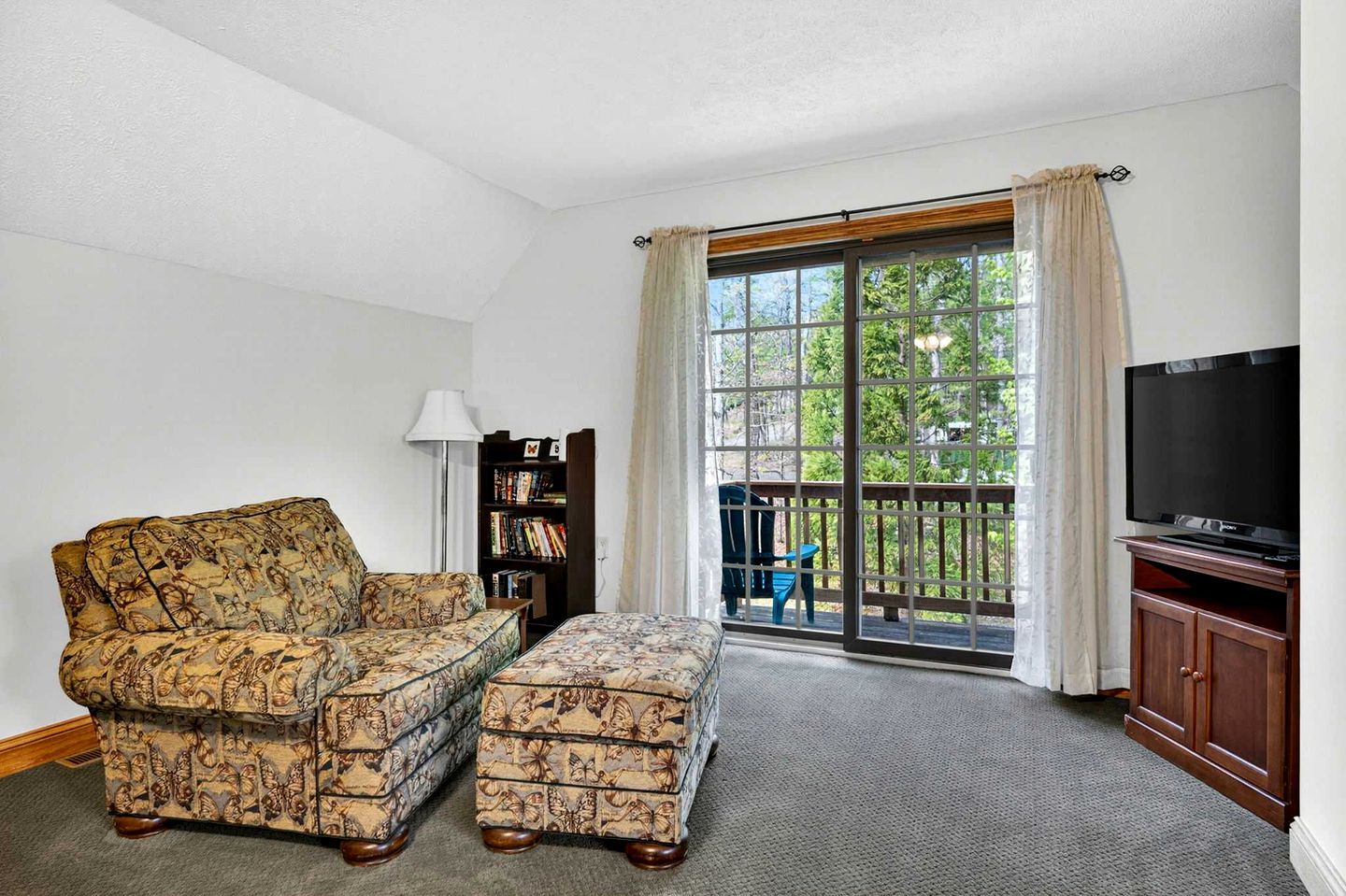 Ein Wohnzimmer mit Sofa, Hocker, TV-Schrank, Bücherregal und einer Schiebetür zu einem Außenbereich.