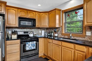 A kitchen with wooden cabinets, a black microwave, a stove, and a window with a view of trees. - Mountain Lodge with Hot Tub & Fire Pit in Berkeley Springs, West Virginia (Berkeley Springs)