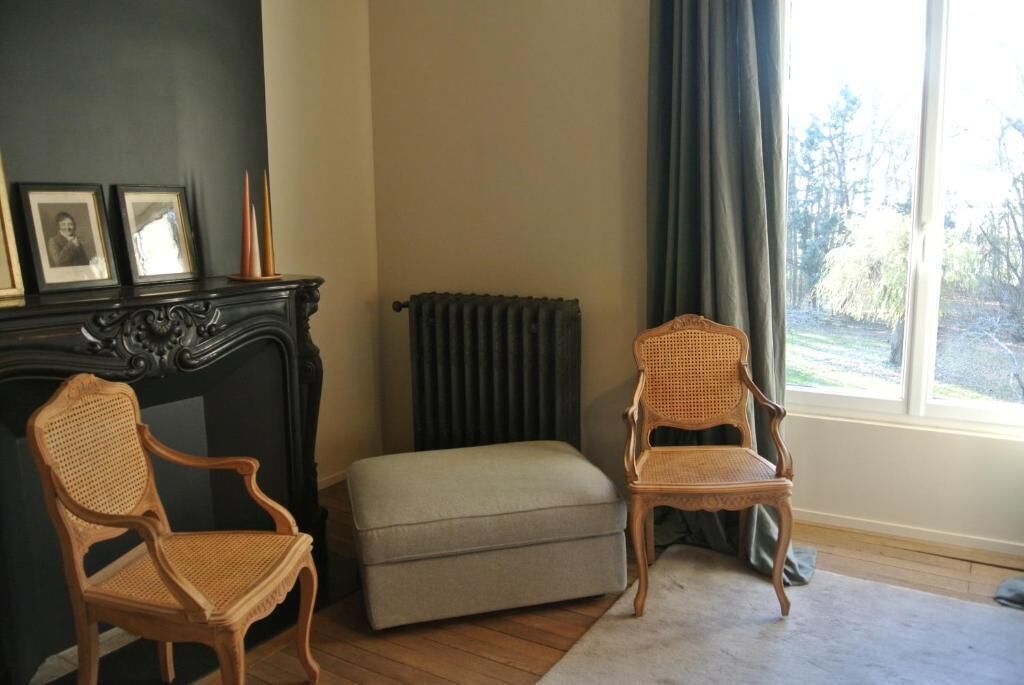 Een kamer met twee rieten stoelen, een klein poufje, een open haard met ingelijste foto's en uitzicht door een raam.