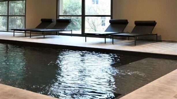 An indoor swimming pool with clear windows, lounge chairs, and a tiled floor.