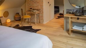 A bedroom with a sloped ceiling, exposed wooden beams, a brick wall, a wooden desk, a chair, a bed, a round mirror, and a wooden vanity with a bowl.