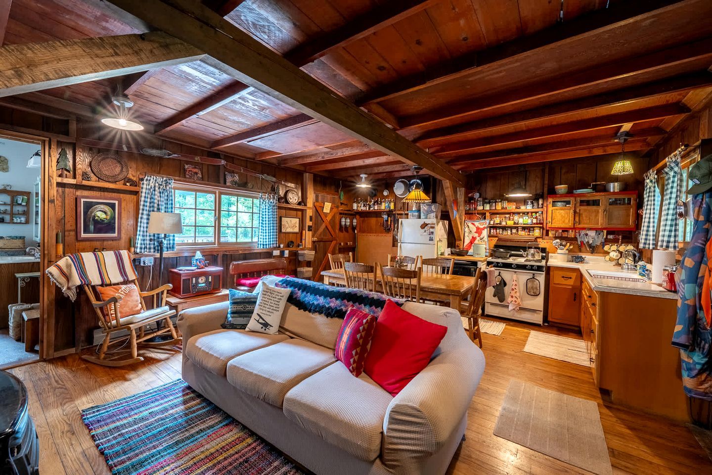 Interior acolhedor de uma cabana com zona de refeições em madeira, cozinha com fogão e sala de estar com sofá e poltrona de balanço.