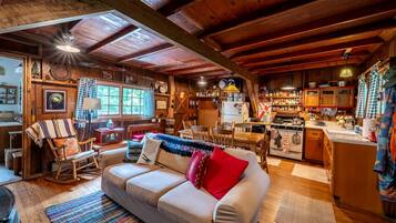 Interior acolhedor de uma cabana com zona de refeições em madeira, cozinha com fogão e sala de estar com sofá e poltrona de balanço.