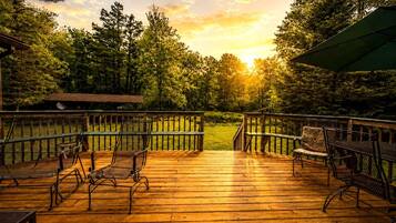 Um deck de madeira com cadeiras e uma mesa de metal, cercado por um muro de madeira e árvores, com um pôr do sol ao fundo.