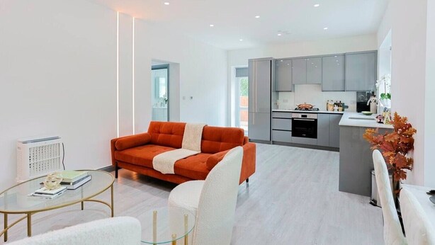 A modern living room with a sofa, a glass coffee table, and a white armchair. There is a kitchen area with grey cabinets and a white countertop.