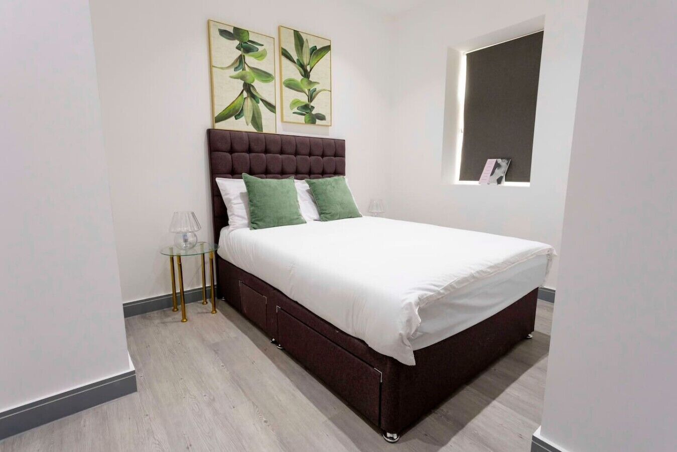 A bedroom with a bed, a nightstand, a window, and two wall-mounted paintings.