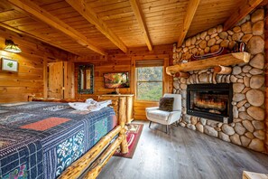 Ein Blockhaus-Interieur mit Kamin aus Stein, Holzwänden, einem Bett mit gemustertem Überwurf, einem Fernseher und einem Fenster mit Blick.