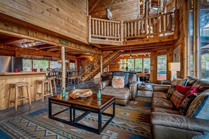 Interior d'una cabana de fusta amb zona de menjador, sala d'estar amb sofà i taula de centre, i escala que mena a una planta superior.