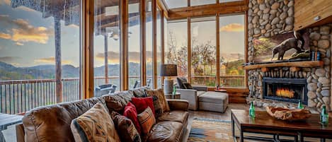 A spacious living room with a leather sofa, a stone fireplace, and large windows offering a mountain view.