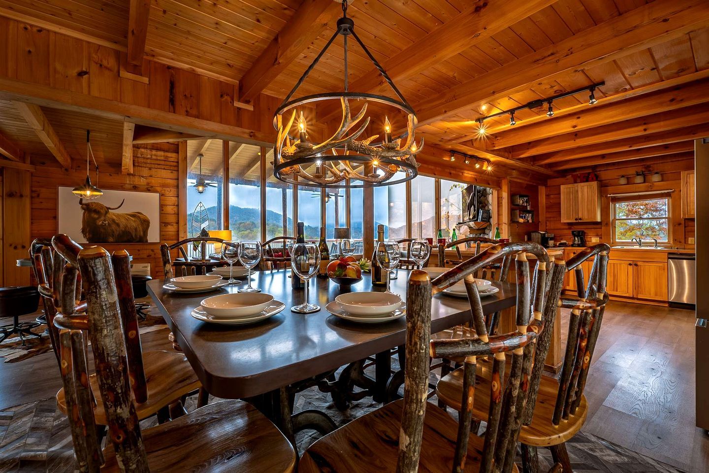 Un intérieur en bois avec une table à manger dressée pour quatre personnes, entourée de chaises rustiques, d’un lustre et d’une grande fenêtre offrant une vue sur les montagnes.