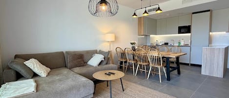En moderne stue med sofa, spisebord med stole, et rundt bord med fjernbetjeninger og et køkken i baggrunden.