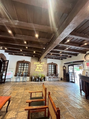 A spacious room with a high ceiling, stone flooring, and wooden beams. There are several chairs and tables, a clock, and a sign indicating an exit.