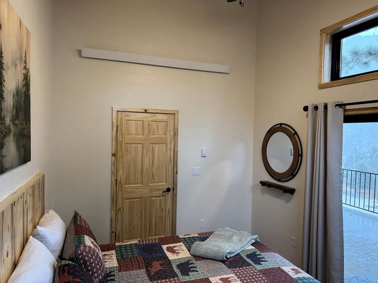 Un dormitorio con puerta de madera, colcha acolchada, espejo redondo y ventana con vistas a árboles.