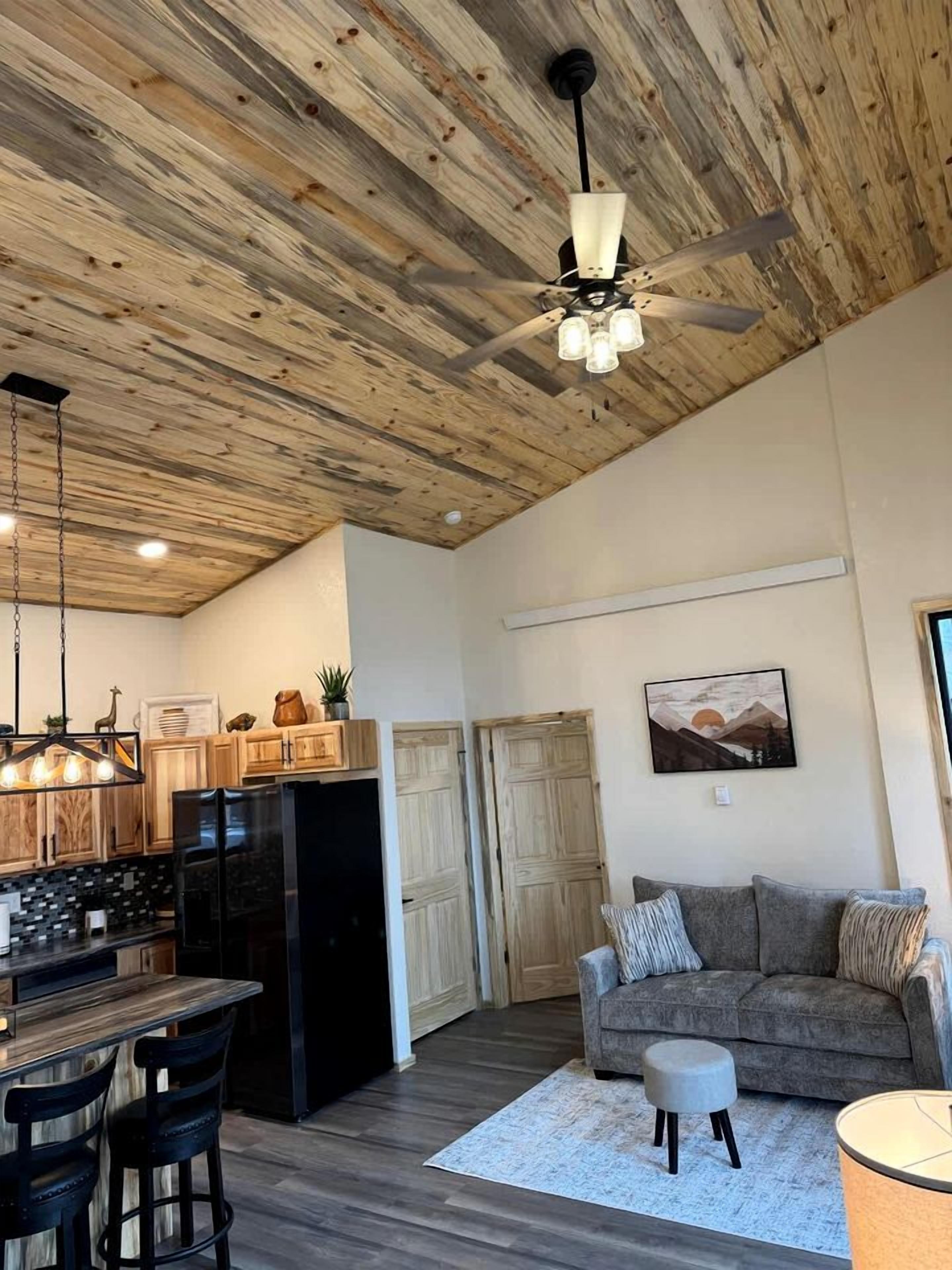 Amplia sala de estar con techo de madera, comedor con mesa y sillas de madera, refrigerador negro y sofá gris con un pequeño otoman.