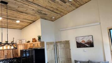 Amplia sala de estar con techo de madera, comedor con mesa y sillas de madera, refrigerador negro y sofá gris con un pequeño otoman.