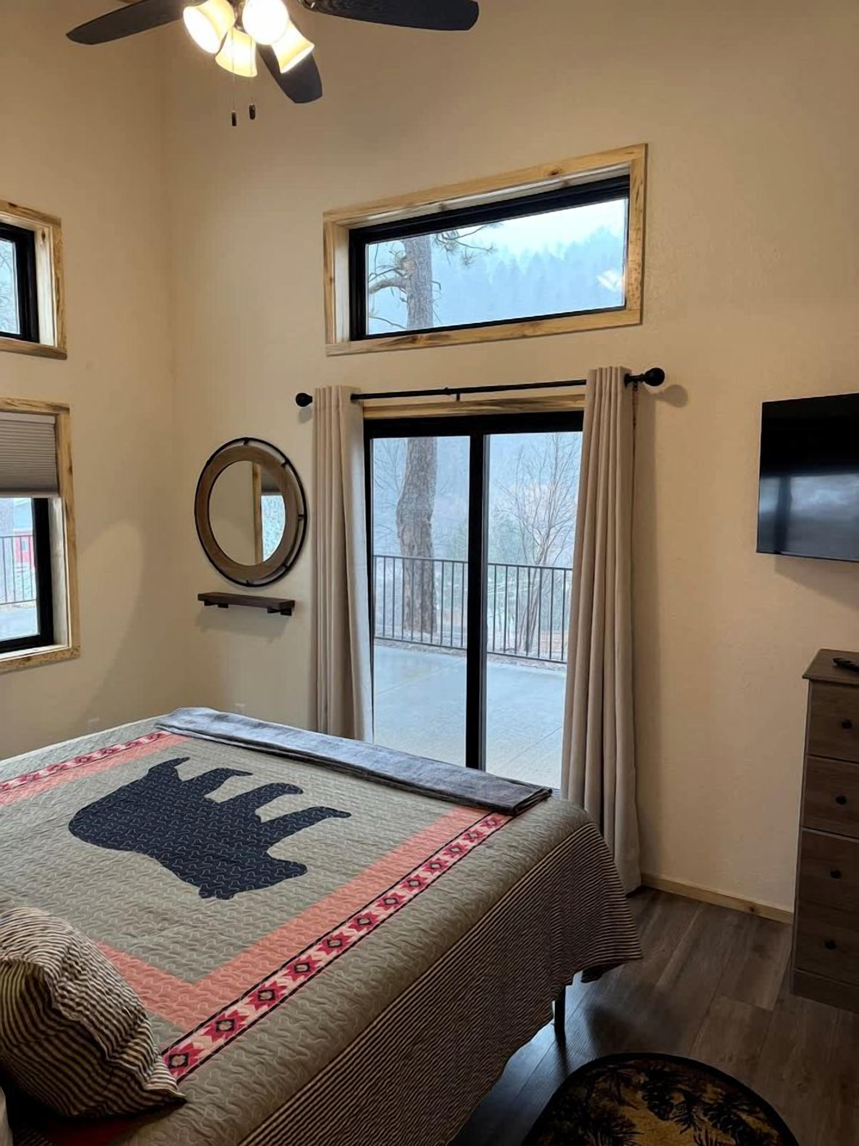 Un dormitorio con una cama, ventilador de techo, ventana grande, televisor y vista a un balcón.