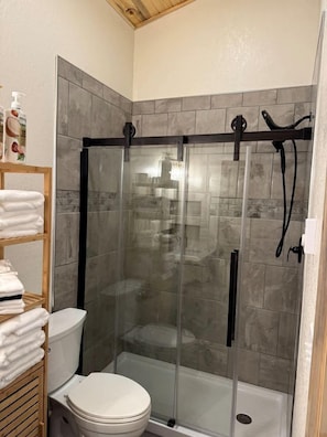 A shower enclosure with a glass door, a toilet, and a shelf with folded towels. - Radiant Family Vacation Rental Retreat near Black Hills Mountain in South Dakota (Keystone)