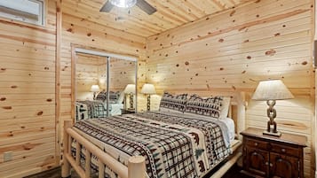 Un dormitorio de cabaña de madera con una cama, dos mesitas de noche con lámparas, un ventilador de techo y un espejo grande.