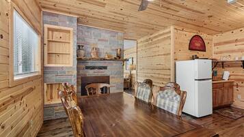 A wooden cabin interior with a stone fireplace, wooden dining table, chairs, a white refrigerator, and a ceiling fan.