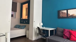 A compact room with a bed, a desk, and a chair, a blue accent wall, and a framed picture of a cityscape.
