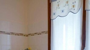Un baño con pared de azulejos blancos, estante de madera con toallas, radiador y ventana con cortina.