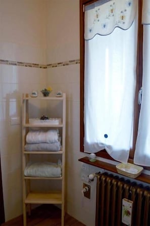 A bathroom with a white tiled wall, a wooden shelf with towels, a radiator, and a window with a curtain.