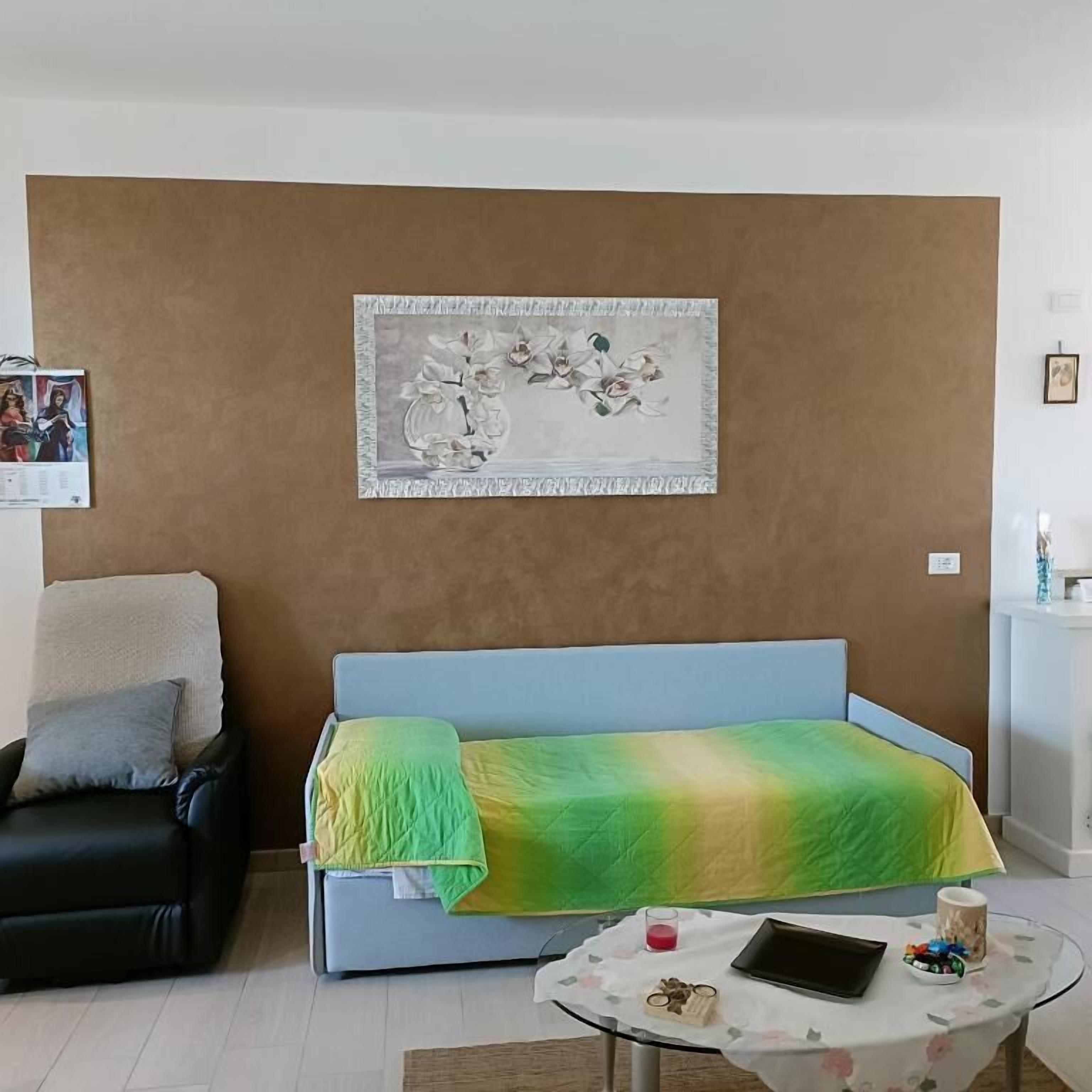 Una habitación con un sofá azul, una pequeña mesa redonda con una tableta, una obra de arte enmarcada en la pared y una silla negra.