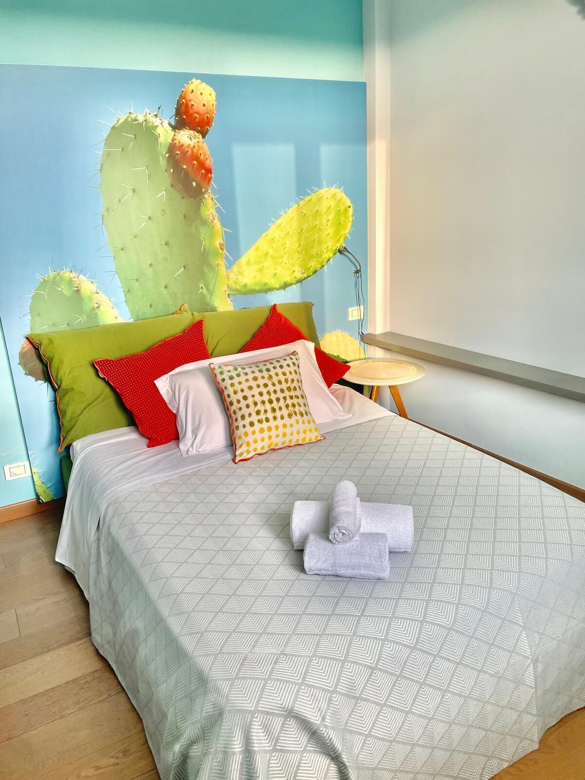 A bed with a geometric patterned bedspread, pillows in various colors, a cactus decoration, and a small bedside table with a lamp.