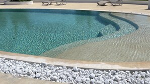 A clear pool with lounge chairs, surrounded by a pebble border, and trees in the background. - Masseria Sant'Eleuterio (Collepasso)