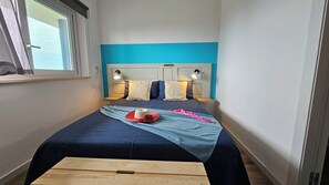 Ein Bett mit blauer Kopfteil, zwei Nachttischlampen und eine hölzerne Sitzbank in einem Zimmer mit Fenster.