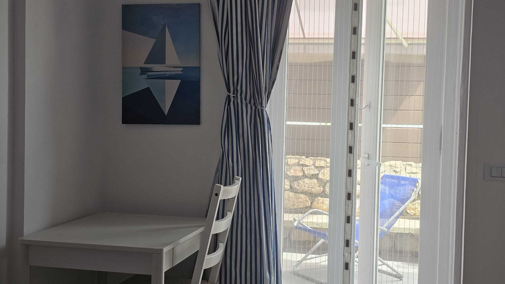 Una stanza con una sedia bianca, una tenda a righe, un quadro raffigurante una barca a vela e vista su un muro di pietra e sedie blu all'esterno.