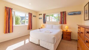 A bedroom with a large bed, two bedside tables with lamps, a dresser, and two windows with curtains. - Farrant Family Escape – Roomy Wanaka Retreat (Wānaka)