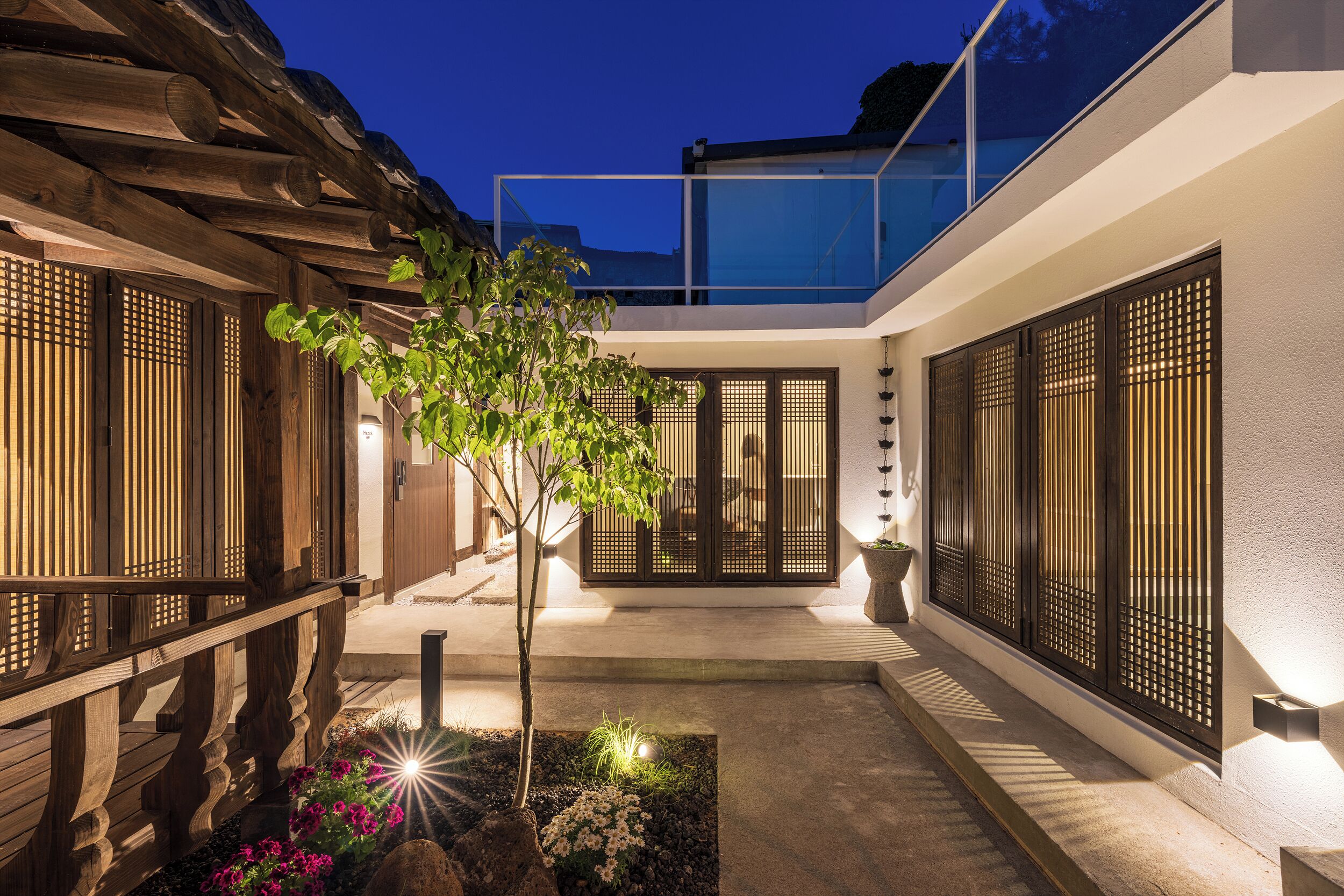 A traditional Japanese-style building with a wooden exterior, a small garden with a tree and flowers, and a modern glass extension.
