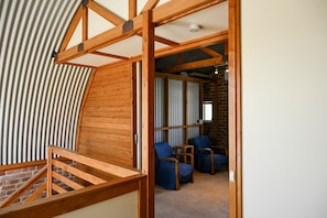 Habitación estilo loft con vigas de madera a la vista, techo inclinado y pared de ladrillo. Hay dos sillas azules y una mesita.