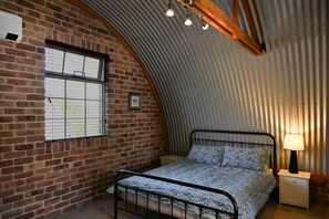 Une chambre avec un mur en briques, un cadre de lit en métal, une table de chevet avec une lampe et une fenêtre avec des stores.