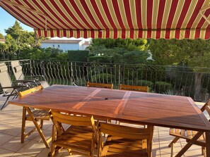Eine Terrasse im Freien mit Holztisch und -stühlen, einem Streifen-Markisen und Blick auf einen Garten und Gebäude.