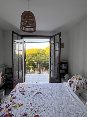 A bedroom with a bed, a chair, a bedside table, a lamp, and a bookshelf. There is an open door leading to a balcony with a view of trees and a fence. - Beachfront family villa with a pool, Domaine de Port d'Alon, sleeps 6–10 (Saint-Cyr-sur-Mer)