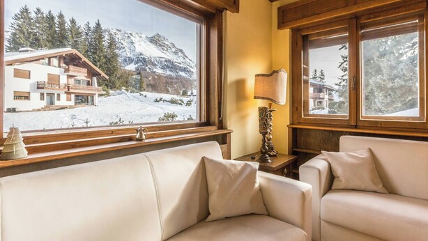 Una stanza accogliente con una grande finestra che si affaccia su un paesaggio montano innevato, un soffitto in legno e un divano in pelle bianca.
