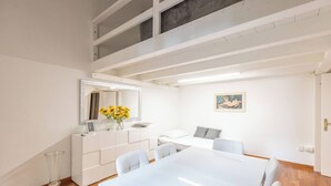 Une pièce moderne avec une grande table à manger blanche, des chaises blanches, un canapé blanc et un grand miroir agrémenté d’un vase de fleurs jaunes.