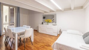 Une pièce moderne avec une table à manger blanche, des chaises, un canapé blanc, un plancher en bois, un meuble blanc et une petite fenêtre avec des rideaux.