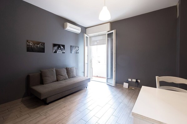 Un salon moderne avec un canapé, une table à manger et une chaise. On peut voir des unités de climatisation fixées au mur, ainsi qu’une porte donnant sur l’extérieur.