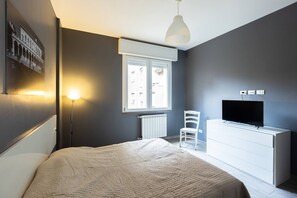 Une chambre à coucher comprenant un lit, une commode blanche, un téléviseur, une chaise, une fenêtre avec des stores, un luminaire fixé au mur et un radiateur.