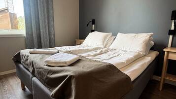 Un lit recouvert d’une couverture marron, avec des oreillers blancs et une serviette pliée posée dessus. Il y a une fenêtre avec des rideaux, une table de chevet avec une lampe et un plancher en bois.