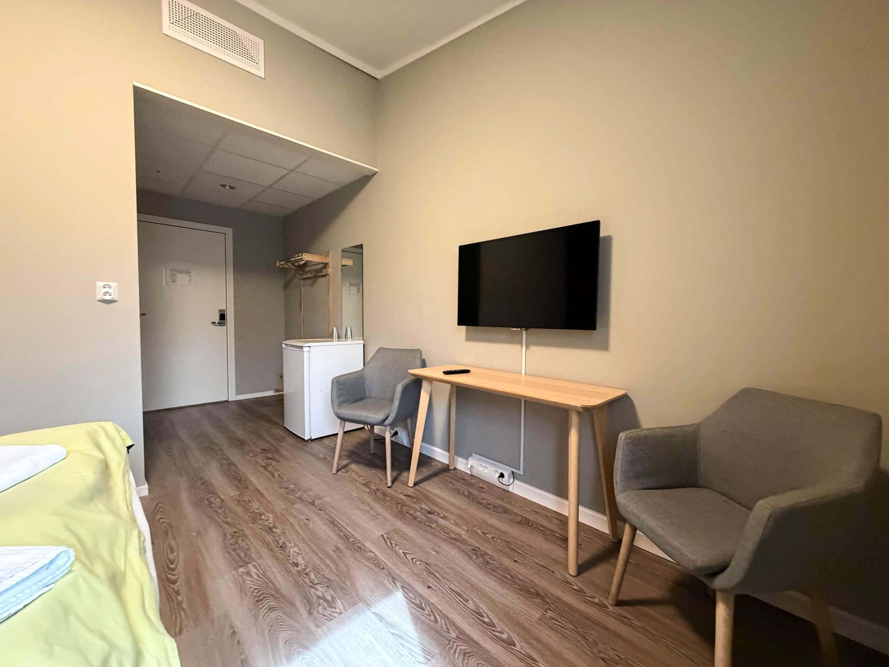A room with a wooden floor, a desk with a chair, a television on the wall, and a bed with a green cover.