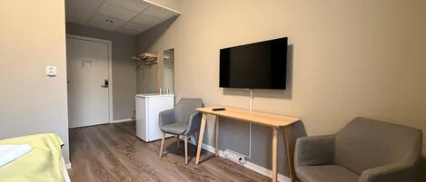A room with a wooden floor, a desk with a chair, a television on the wall, and a bed with a green cover.