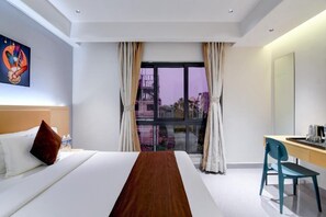 A hotel room with a bed, a desk, a chair, a window with curtains, and a painting on the wall.